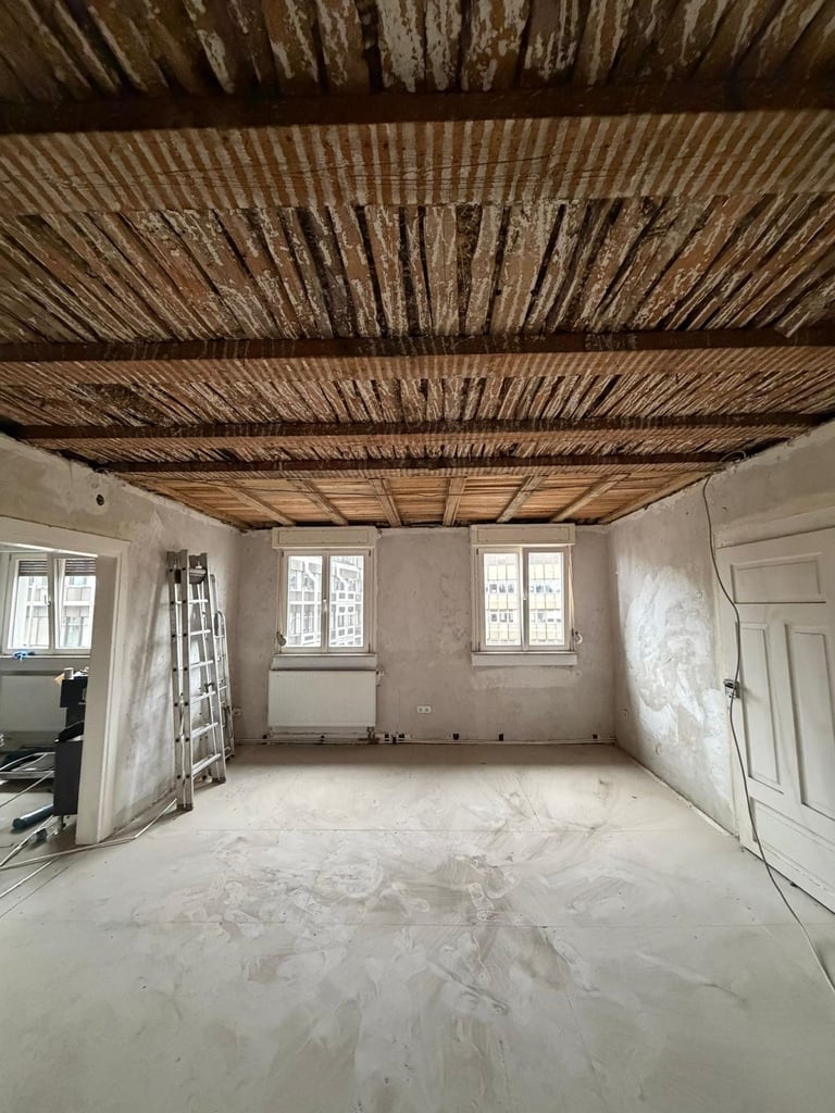 Leerer Raum mit Holzbalken, weißen Wänden und Betonboden vor der Renovierung