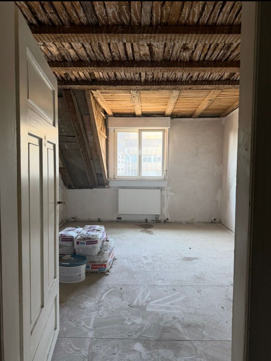 Raum während Renovierung mit Holzbalkendecke, weißen Wänden, Fenster mit Heizkörper und Baustoffen auf staubigem Boden